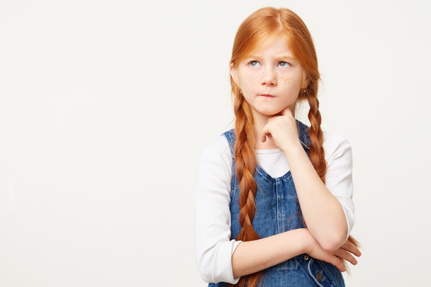 Μικρό παιδί κόκκινα μαλλιά κορίτσι με δύο μακριές πλεξούδες σκέφτεται, φαίνεται στην αριστερή άνω γωνία δύσπιστα με αμφιβολία, μια γωνία των χειλιών στιγκάρει, κρατά γροθιά κάτω από το πηγούνι, άσπρο φόντο, Τζιν συνολική - Φωτογραφία, εικόνα