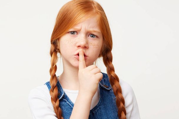 Close up of serious worried red-haired girl depicts silence put her index finger to lips, isolated on a white background. - Photo, Image