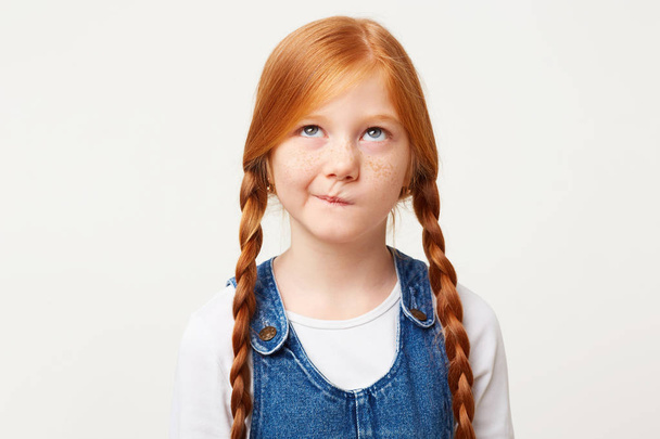 Serious little girl with freckles and braided in two long plaits red hair thinks, looks up incredulously, ponders about kindergarten, with doubt, one corner of the lips pursed, white background - Photo, Image