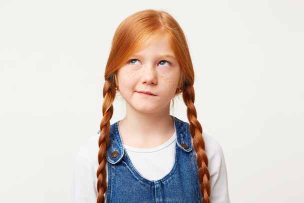 Little girl with freckles and braided in two long plaits red hair thinks, looks aside incredulously, with doubt, one corner of the lips pursed, white background, dressed in denim overall - Photo, Image