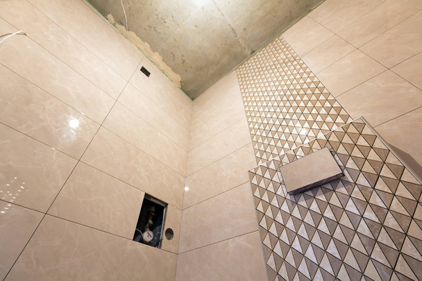 Unfinished reconstruction of bathroom or toilet with light beige geometric mosaic ceramic tiles installed on walls, place for lavatory and bath. Low angle, bottom view. - Photo, Image
