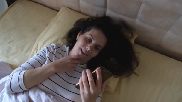 funny woman laughs take a picture. Portrait of a caucasian young woman lying in bed with a millennial pillow - Imágenes, Vídeo