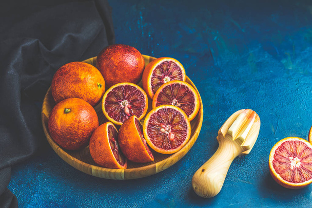 Φέτες και ολόκληρα φρούτα Σικελίας αίμα πορτοκάλια σε ξύλινη πινακίδα και αποχυμωτή πάνω από σκούρο μπλε συγκεκριμένο πίνακα επιφάνεια. Σκοτεινό ρουστίκ στυλ. - Φωτογραφία, εικόνα