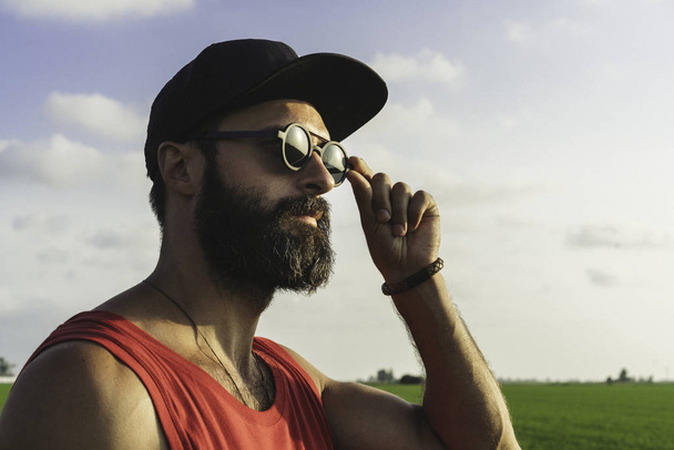 Bearded man model wearing black cap, sunglasses and red tshirt looks away, sunset in the green rice fields scenery - Photo, image