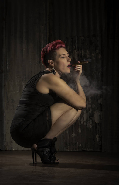 Sultry woman with fuchsia hair crouching and holding a cigar - Fotoğraf, Görsel
