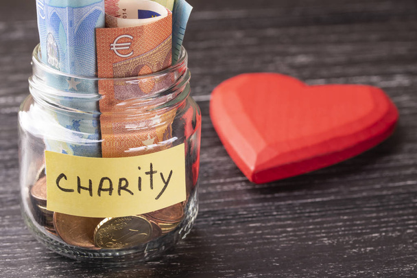 Glasgefäß mit Münzen und Euro-Scheinen mit der Aufschrift Charity. mit einem riesigen roten Herz auf einem alten schwarzen Holztisch. das Konzept der Güte und Hilfe für Bedürftige. - Foto, Bild