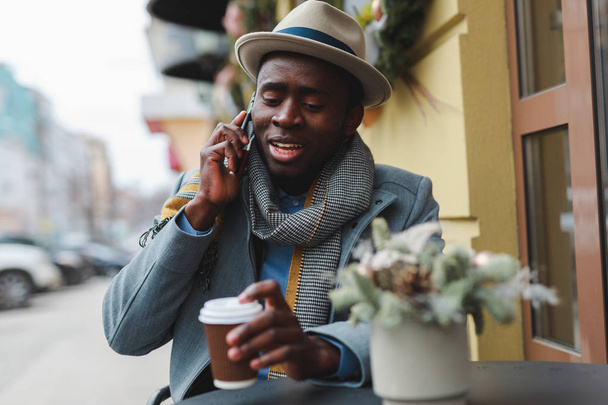 Happy african american man talking with friend by smartphone sitting at outdoor cafe - Foto, imagen