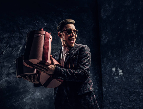 Κομψά ντυμένος άντρας κρατώντας δώρα ενώ θέτοντας σε ένα σκοτεινό στούντιο - Φωτογραφία, εικόνα