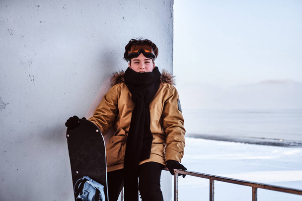 ein junger Snowboarder in warmer Kleidung und Schutzbrille sitzt auf einem Geländer in der Nähe des verschneiten Strandes - Foto, Bild