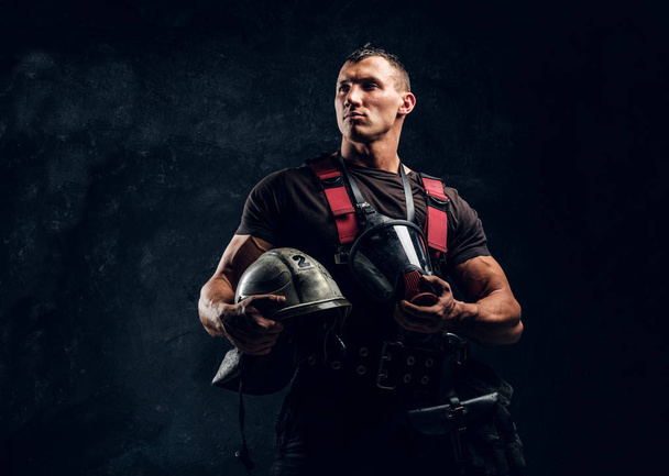 Handsome muscular fireman holding a helmet and oxygen mask standing in the studio against a dark textured wall - Photo, image