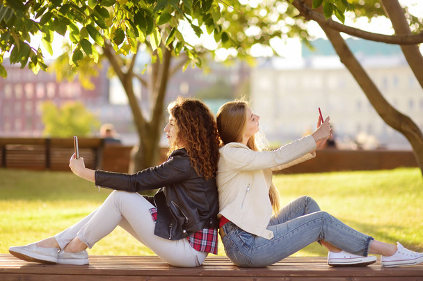 Two beautiful young women take selfies in a sunny park at the same time. Girlfriends. Communication. Always in touch. - Foto, imagen