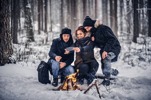 Group of cheerful friends travelers make Sefley sitting by the campfire in the midst of a snowy forest - Photo, image