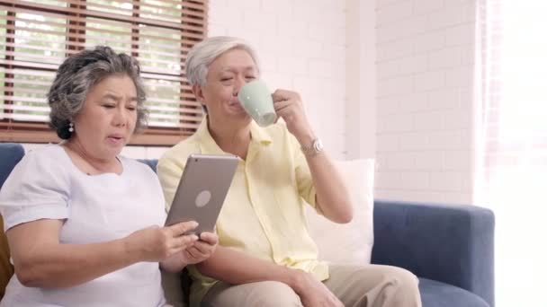 Casal de idosos asiáticos usando tablet e beber café na sala de estar em casa, casal desfrutar de momento de amor enquanto deitado no sofá quando relaxado em casa. Desfrutando de tempo estilo de vida família sênior em casa conceito
. - Filmagem, Vídeo