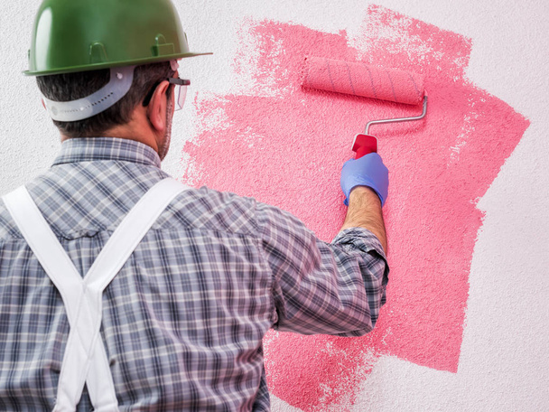 Pintor de casa caucásico trabajador en overoles de trabajo blanco, con el rodillo pinta la pared con la pintura de color rosa. Industria de la construcción. Seguridad en el trabajo
.  - Foto, Imagen