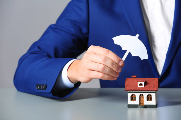Man covering house model with umbrella cutout at table, closeup. Home insurance - Photo, image