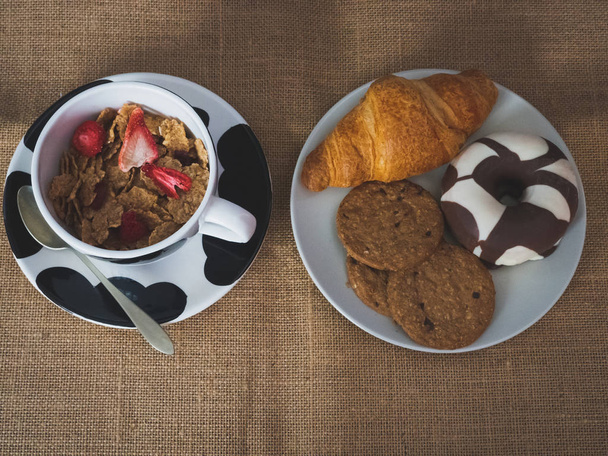 Breakfast of a porcelain bowl with cereals with red berries, an antique spoon and one plate with one croissant, one chocolate donut and cookies on a vintage burlap background - Zdjęcie, obraz