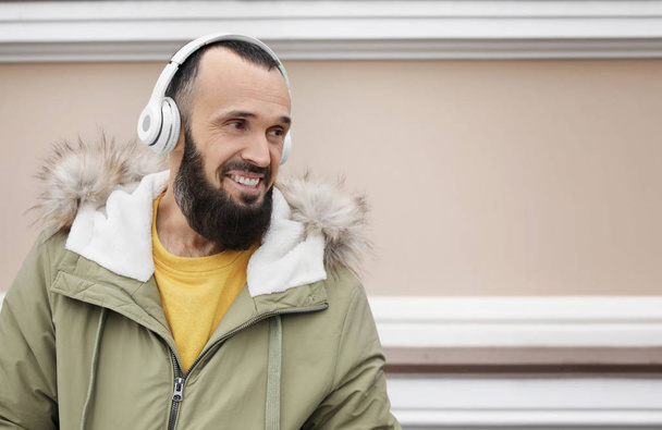 Mature man with headphones listening to music near light wall. Space for text - Фото, изображение