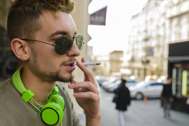 ストリートでタバコを楽しんで緑のヘッドフォンとサングラスを持つ若いハンサムな男. - 写真・画像