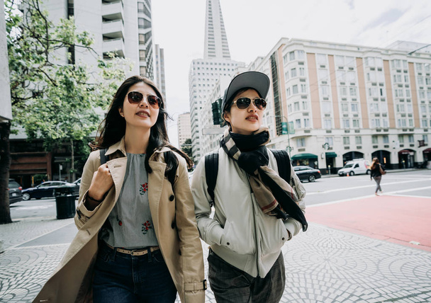 two asian college girls going to university together walking in front ground of university school in busy area san francisco. group of sisters in sunglasses standing with transamerica pyramid in back - Фото, изображение