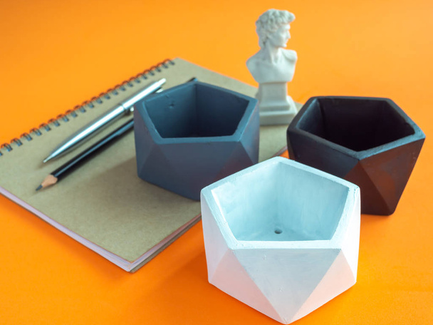 Modern geometriai pentagon beton virágtartók, jegyzetfüzet, toll, ceruza, és kis Dávid szobor, narancsszínű háttér. Festett beton cserepek lakberendezési minimalista stílus. - Fotó, kép