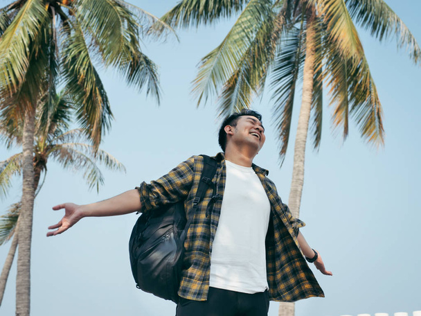 Asian man with backpack raising his hands with blissful feeling and coconut tree background, lifestyle concep.t - Foto, afbeelding