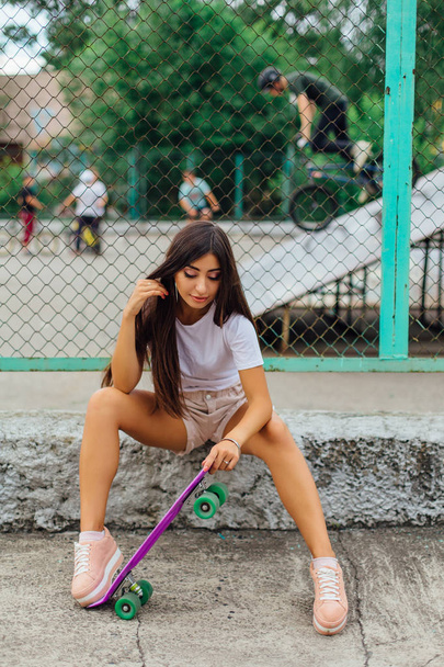 Summer lifestyle image of trendy pretty young girl sitting next to the skateboard coart with her purple plastic skateboard. - Fotoğraf, Görsel