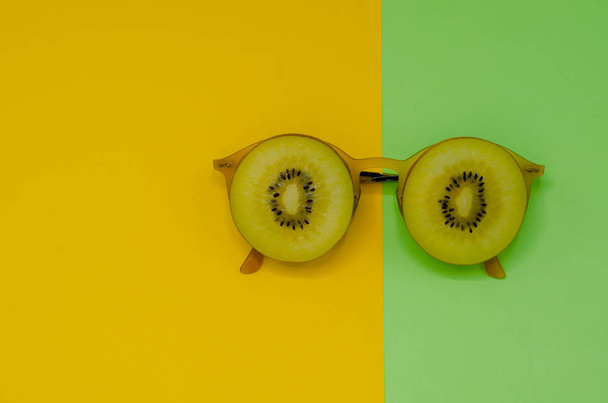 Two slice golden kiwi fruits put on yellow sunglasses set as face with eyes photo isolated on yellow and green background for summer time with space for text. - Photo, Image