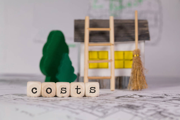 Слово COSTS состоит из деревянной буквы. Небольшой бумажный дом, деревянные деревья на заднем плане. Крупный план
 - Фото, изображение
