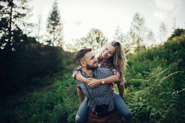Молодий веселий чоловік тримає дівчину на свинарстві назад у зеленому парку на заході сонця
  - Фото, зображення