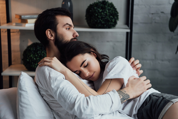 attractive young tattooed couple embracing while lying in bed at home - Fotoğraf, Görsel