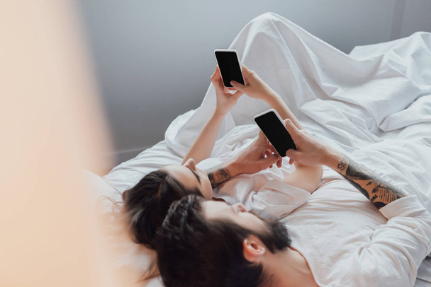 νεαρό ζευγάρι ξαπλωμένη στο κρεβάτι και χρησιμοποιώντας τα smartphones με την κενή οθόνη - Φωτογραφία, εικόνα