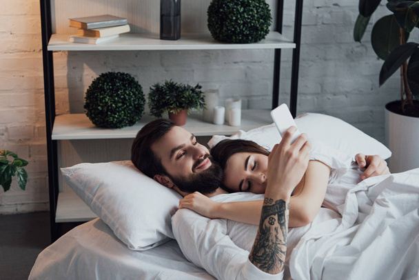 handsome man using smartphone while woman sleeping in bed - Foto, afbeelding
