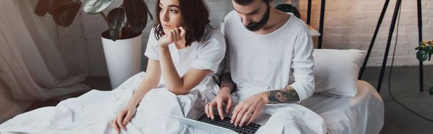 bearded man lying in bed and using laptop while bored woman propping chin in bedroom - Photo, Image
