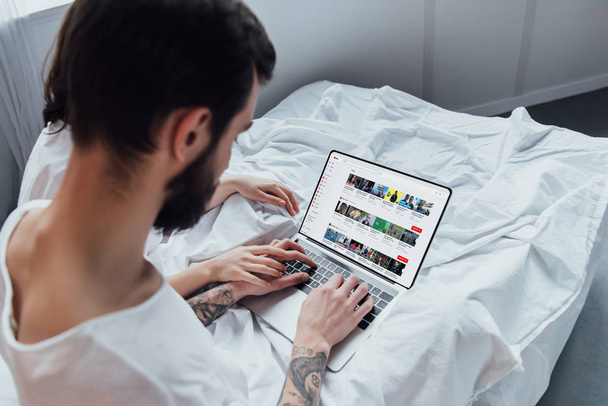 Rückansicht des Paares, das im Bett liegt, Hände hält und Laptop mit youtube-Webseite auf dem Bildschirm benutzt  - Foto, Bild