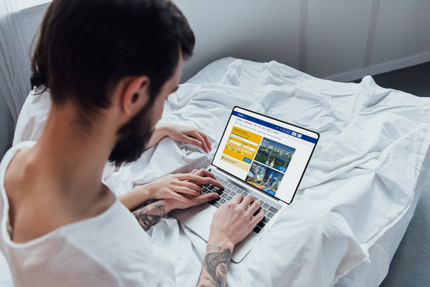 back view of couple lying in bed, holding hands and using laptop with booking website on screen  - Zdjęcie, obraz