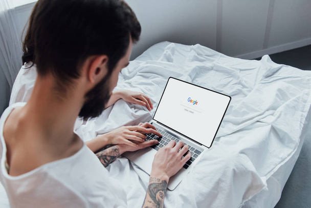 back view of couple lying in bed, holding hands and using laptop with google website on screen  - Fotó, kép