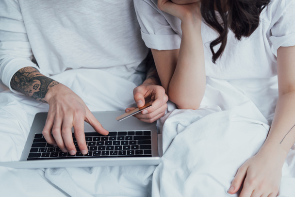 обрезанный вид татуированной пары с кредитной картой и ноутбуком, лежащих в постели и делать онлайн-покупки
 - Фото, изображение