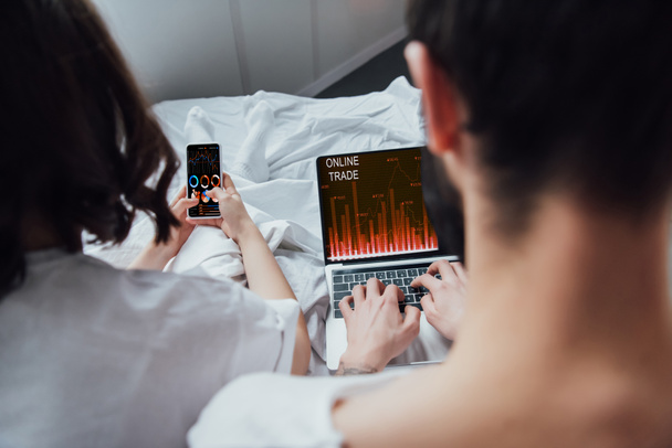 Achteraanzicht van paar liggend in bed en het gebruik van digitale apparaten met online handel en grafieken op scherm  - Foto, afbeelding