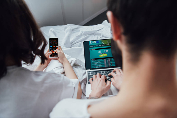 πίσω όψη του ζευγάρι ξαπλωμένη στο κρεβάτι και χρησιμοποιώντας ψηφιακές συσκευές με sportsbet και γραφήματα στην οθόνη  - Φωτογραφία, εικόνα