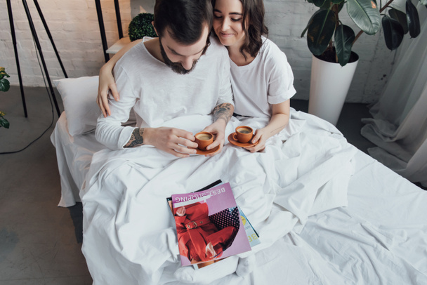 όμορφο νεαρό ζευγάρι ξαπλωμένη στο κρεβάτι με φλιτζάνια καφέ και αγκαλιάζει πρωί - Φωτογραφία, εικόνα