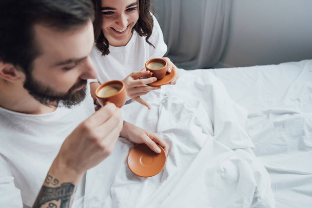 όμορφο νεαρό ζευγάρι πίνοντας καφέ ενώ ξαπλωμένη στο κρεβάτι πρωί - Φωτογραφία, εικόνα
