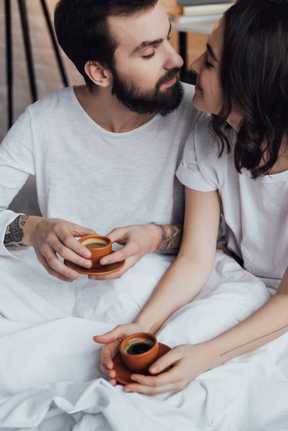 beautiful young tattooed couple with coffee lying in bed and and looking at each other - Foto, afbeelding