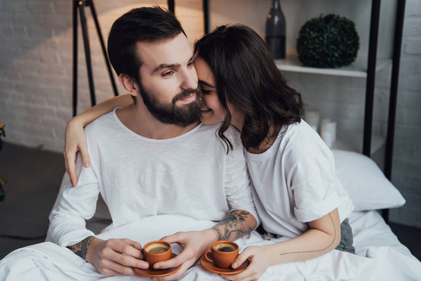 mooie jonge koppel liggend in bed met koffie cups en knuffelen in ochtend - Foto, afbeelding