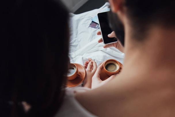 自宅に空白の画面を持つスマート フォンを使用してコーヒー カップを持つカップルの背面します。 - 写真・画像
