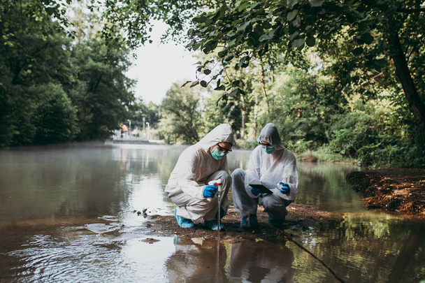 Koruyucu giysili iki bilim adamı nehirden su örnekleri alıyor.. - Fotoğraf, Görsel