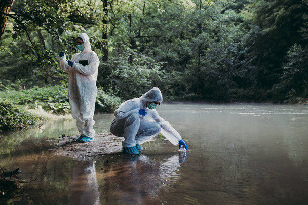 Two scientists in protective suits taking water samples from the river - Фото, изображение