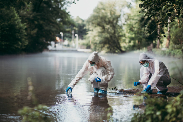 Два учёных в защитных костюмах берут образцы воды из реки
. - Фото, изображение