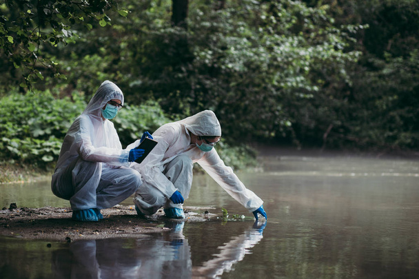 Koruyucu giysili iki bilim adamı nehirden su örnekleri alıyor. - Fotoğraf, Görsel