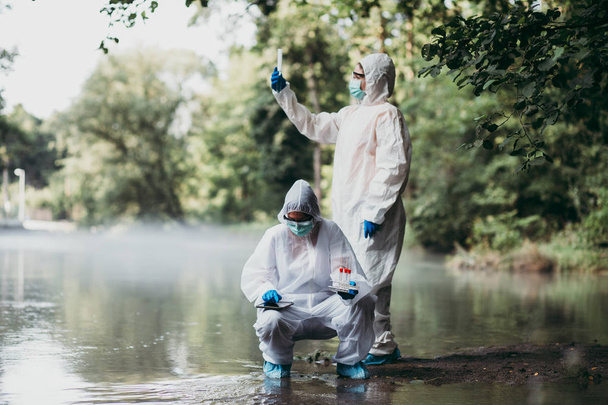 Two scientists in protective suits taking water samples from the river. - 写真・画像
