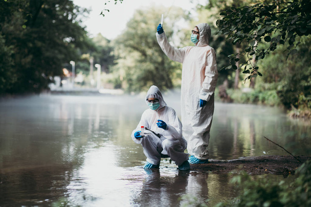 Δύο επιστήμονες με προστατευτικές στολές που παίρνουν δείγματα νερού από το ποτάμι. - Φωτογραφία, εικόνα